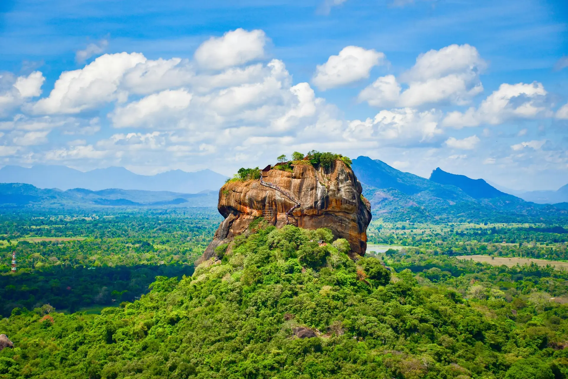 Klippfästningen Sigiriya reser sig över grön skog, med berg vid horisonten